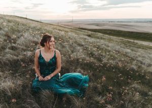 Smiling woman in teal sequin dress sitting in a wildflower meadow at sunset. Senior grad photos. Graduation photos.