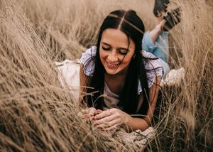 Smiling dark-haired woman lying in golden wheat field during outdoor portrait session. Senior grad photos. Graduation photos.