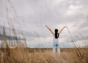 Woman with arms raised standing in golden prairie field under dramatic cloudy sky. Senior grad photos. Graduation photos.