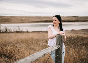 Dark-haired woman in white top leaning on wooden fence post beside a prairie lake. Senior grad photos. Graduation photos.
