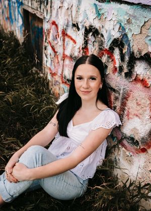 Young woman with dark hair in white top and jeans sitting against graffiti wall outdoors. Senior grad photos. Graduation photos.