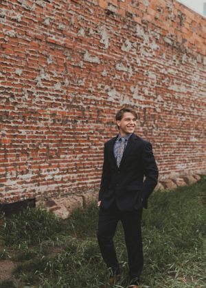Smiling young man in black suit and floral tie stands before weathered brick wall outdoors. Senior grad photos. Graduation photos.