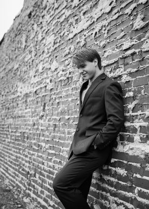 Young man in black suit leans against rustic brick wall smiling in black and white portrait. Senior grad photos. Graduation photos.