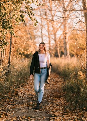 Smiling woman in light jeans and black jacket walking along a leaf-covered autumn forest path. Senior grad photos. Graduation photos.