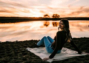 Young woman in jeans sitting on a blanket by a lake shore during a vibrant sunset. Senior grad photos. Graduation photos.