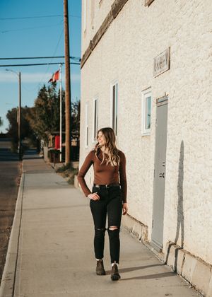 Young woman in brown top and black jeans walking along a sunny small-town sidewalk. Senior grad photos. Graduation photos.