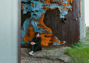 Young man sitting against a rusty metal structure with vibrant peeling blue and orange paint. Senior grad photos. Graduation photos.