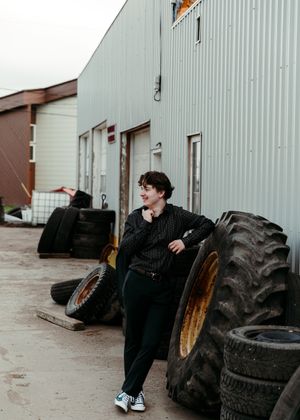Young man leaning against large tractor tires outside an industrial building. Senior grad photos. Graduation photos.