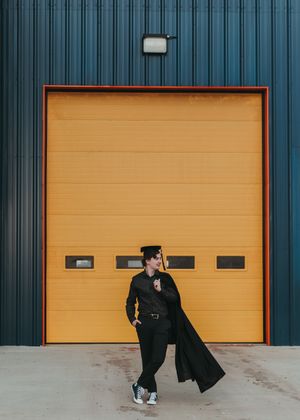Graduate in cap and gown posing in front of a bright yellow garage door. Senior grad photos. Graduation photos.