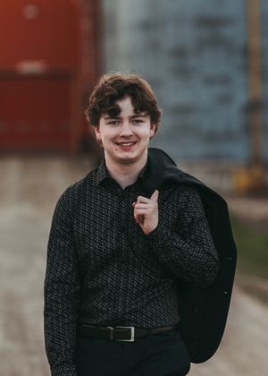 Smiling young man in a dark patterned shirt holding a jacket over his shoulder outdoors. Senior grad photos. Graduation photos.