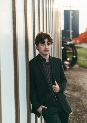 Young man in a dark suit leaning against a metal wall in an industrial setting. Senior grad photos. Graduation photos.