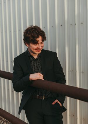 Young man in a black suit leaning on a railing against a corrugated metal wall. Senior grad photos. Graduation photos.