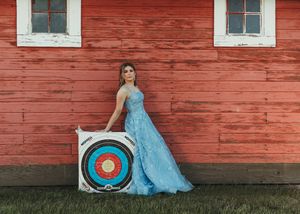 Young woman in blue ball gown posing with archery target beside a red barn wall. Senior grad photos. Graduation photos.