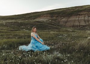 Woman in flowing blue dress sitting in a wildflower meadow with badlands hills behind her. Senior grad photos. Graduation photos.