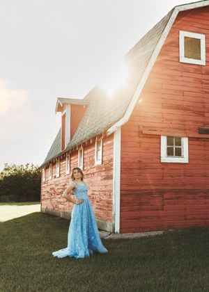 Woman in a blue gown standing beside a classic red barn at sunset. Senior grad photos. Graduation photos.