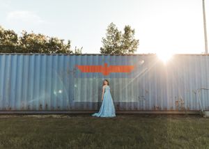 Woman in blue gown standing before a metal shipping container with graffiti in golden sunlight. Senior grad photos. Graduation photos.