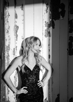 Black and white portrait of blonde woman in sequined black gown posing by a weathered white wall. Senior grad photos. Graduation photos.