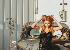 Woman in black sequined gown styles her hair beside a vintage car near a corrugated metal wall. Senior grad photos. Graduation photos.
