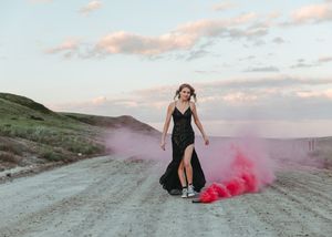 Woman in black gown walks a dirt road surrounded by pink smoke under a dramatic cloudy sky. Senior grad photos. Graduation photos.