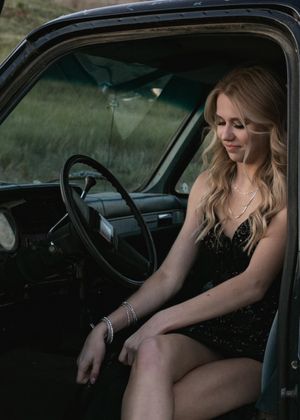 Blonde woman in black sequined mini dress sits inside a vintage truck cab at the steering wheel. Senior grad photos. Graduation photos.
