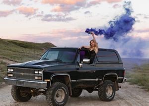Woman holding blue smoke flare from black lifted Chevy Blazer on scenic dirt road at sunset. Senior grad photos. Graduation photos.