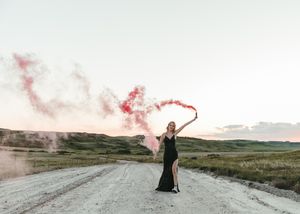 Woman in black gown holding red smoke flare on rural dirt road at golden hour. Senior grad photos. Graduation photos.