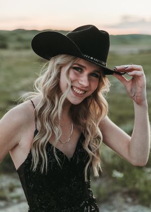 Smiling blonde woman in black sequin dress tipping black cowboy hat outdoors at sunset. Senior grad photos. Graduation photos.