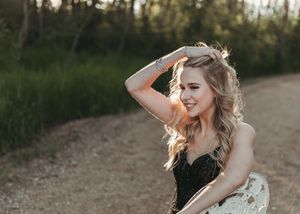 Smiling blonde woman in black lace dress poses outdoors on a dirt path at golden hour. Senior grad photos. Graduation photos.