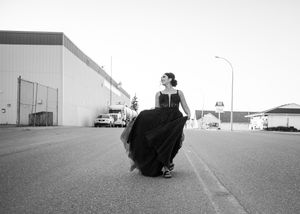Black and white photo of woman in flowing black gown walking across an open road near industrial buildings. Senior grad photos. Graduation p
