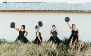 Four women in black ball gowns holding dark bouquets pose in tall grass against a white building. Senior grad photos. Graduation photos.