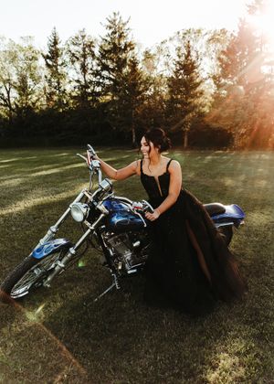 Woman in a black ball gown posing beside a blue motorcycle on a sunlit green lawn. Senior grad photos. Graduation photos.