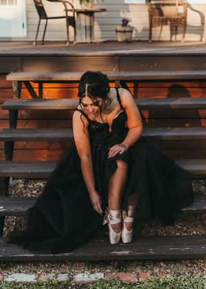 Woman in black tulle gown sits on wooden bleachers lacing up ballet pointe shoes. Senior grad photos. Graduation photos.