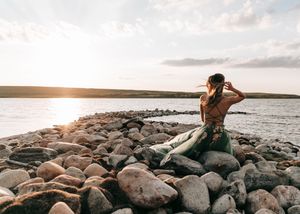 Woman in green gown sitting on rocky lakeside shore gazing at a beautiful sunset. Senior grad photos. Graduation photos.