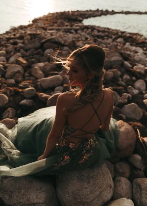 Woman in green floral backless gown sitting on rocky shoreline with wind-blown hair. Senior grad photos. Graduation photos.