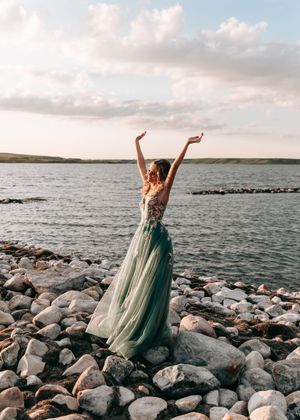 Woman in green floral tulle gown raising arms joyfully on a rocky lakeside at sunset. Senior grad photos. Graduation photos.