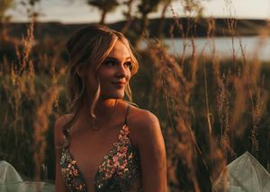 Blonde woman in floral embroidered dress standing in tall golden grass near a lake at dusk. Senior grad photos. Graduation photos.