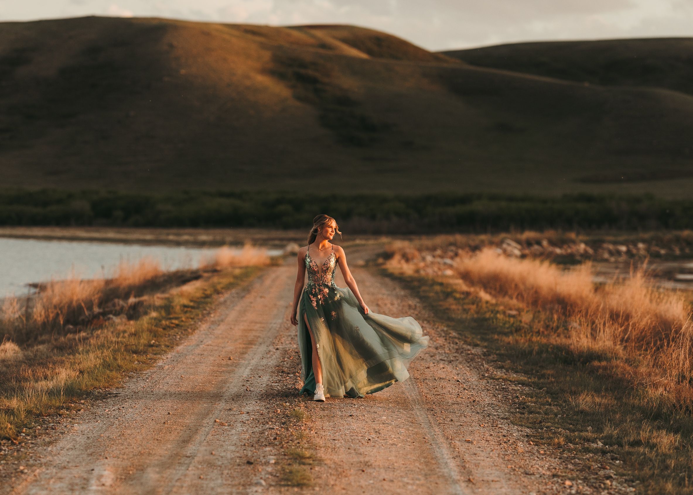 Woman in flowing green gown walks dirt road with scenic hills and lake in golden hour light. Senior grad photos. Graduation photos.