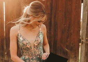 Woman in floral embroidered green gown standing near rustic wooden fence in warm sunlight. Senior grad photos. Graduation photos.