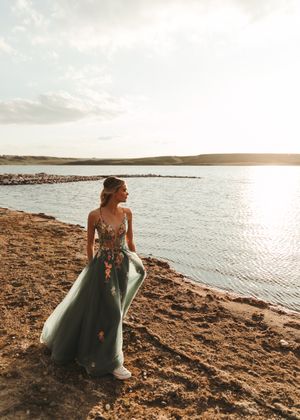 Woman in green floral tulle gown standing by a serene lake shore at golden sunset. Senior grad photos. Graduation photos.