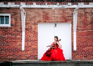 Woman in red ballgown playing cello against a brick wall with white doors. Senior grad photos. Graduation photos.