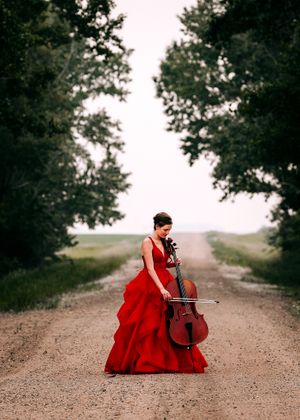 Cellist in a red ballgown playing cello on a scenic tree-lined dirt road. Senior grad photos. Graduation photos.