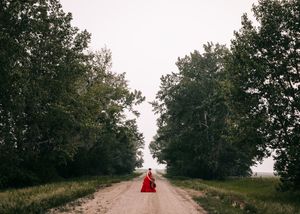 Woman in a red ballgown sitting alone on a tree-lined gravel country road. Senior grad photos. Graduation photos.