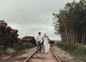 Couple holding hands walking on railroad tracks surrounded by trees and stacked logs under cloudy skies. Senior grad photos. Graduation phot