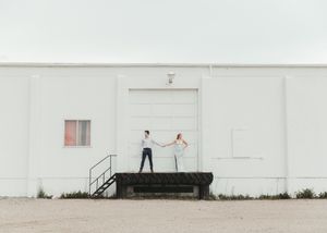 Couple holding hands on a loading dock outside a white industrial building on an overcast day. Senior grad photos. Graduation photos.