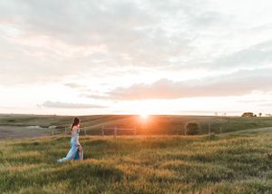 Woman in a blue dress walking through a golden prairie field at sunset with glowing horizon. Senior grad photos. Graduation photos.
