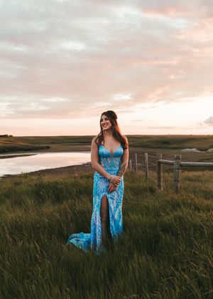 Woman in a blue patterned gown standing in a green field near a pond at sunset. Senior grad photos. Graduation photos.