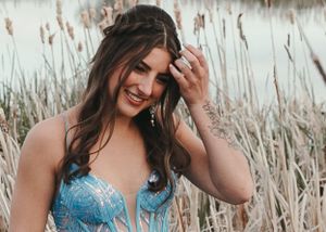 Smiling brunette woman in a blue lace gown touching her hair near tall reeds by a lake. Senior grad photos. Graduation photos.