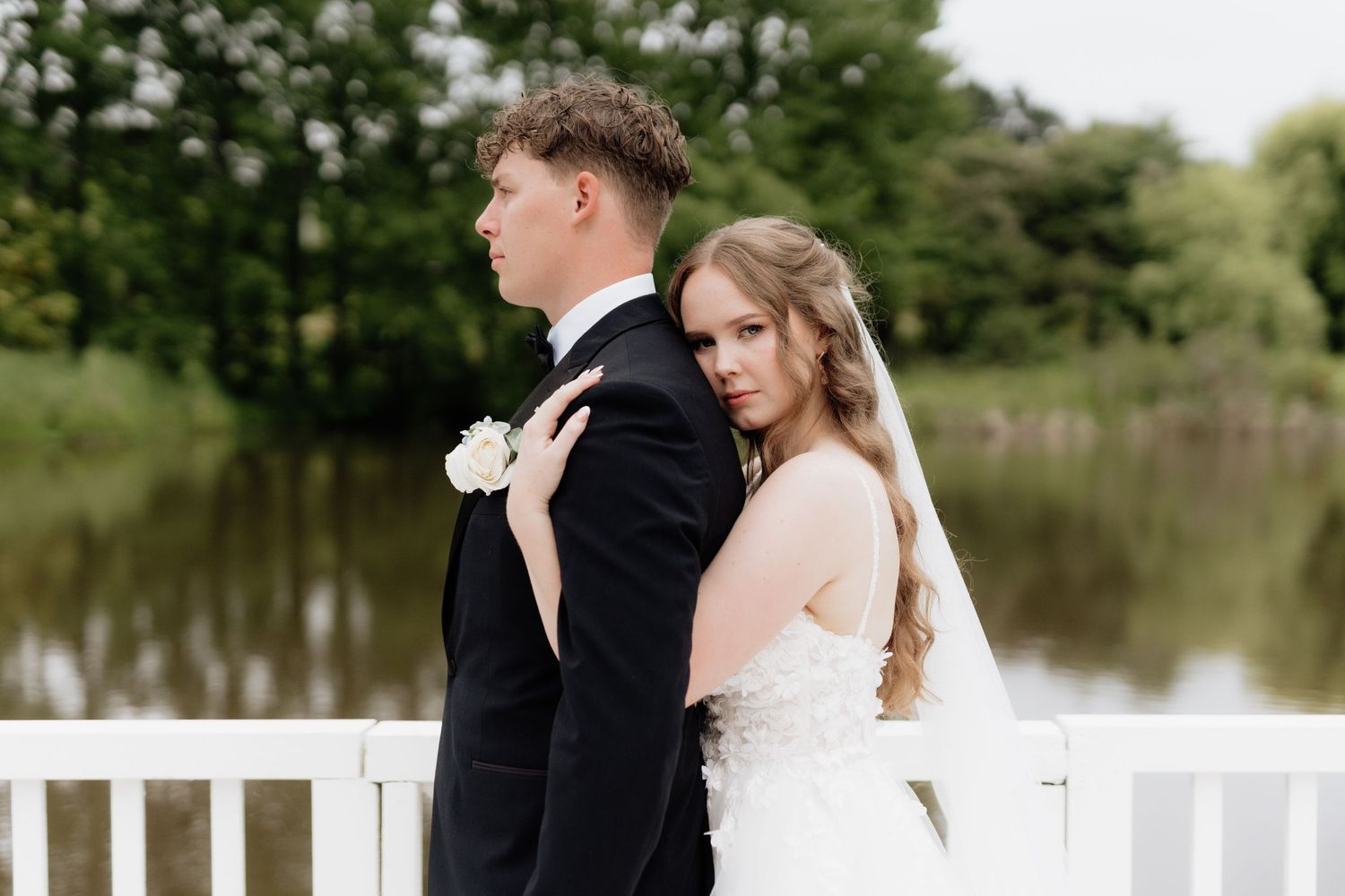 A bride and groom stand back to back on a white deck overlooking a serene lake surrounded by trees.