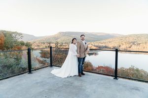 A couple in wedding attire stands on a mountain overlook during sunset, with scenic hills in the background.