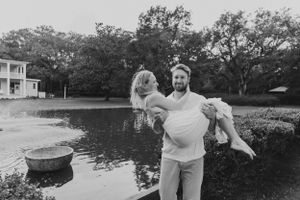 A joyful moment captured in black and white by a lake, with trees and a house visible in the background.